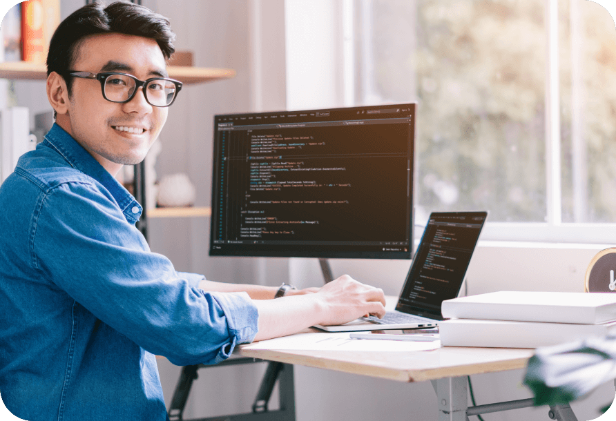 An app developer sits at a desk in front of a laptop and computer monitor with code on the screens.