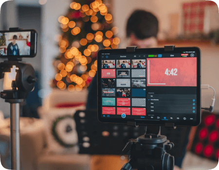 A tablet and smartphone mounted on tripods displaying a digital interface, with a person and a decorated Christmas tree in the blurred background.