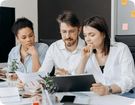 Three individuals are engaged in a work session. The central figure appears to be presenting a document, drawing the attention of the others. The table is strewn with office supplies, suggesting a collaborative and active discussion.